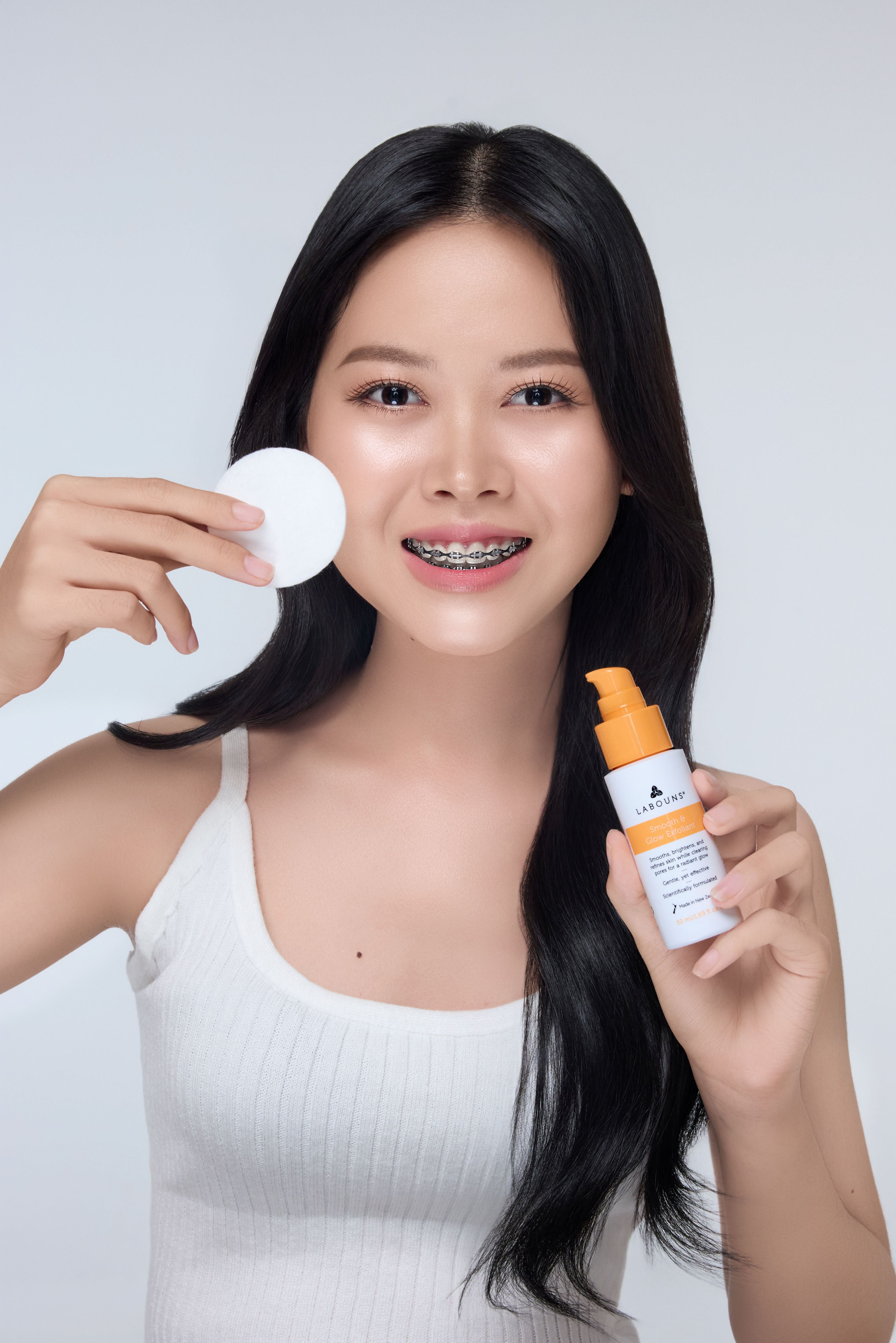 Woman applying facial exfoliant with a bottle labeled 'Labouns Smooth & Glow Exfoliant' and cotton pad on a plain background